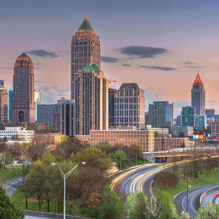 Skyline von Atlanta, USA, bei Sonnenuntergang mit beleuchteten Hochhäusern und fließendem Abendverkehr auf den geschwungenen Highways im Vordergrund.