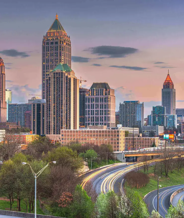 Skyline von Atlanta, USA, bei Sonnenuntergang mit beleuchteten Hochhäusern und fließendem Abendverkehr auf den geschwungenen Highways im Vordergrund.