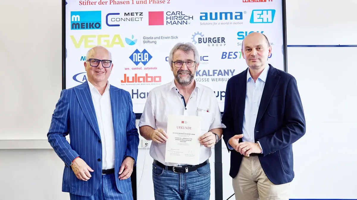 Drei Männer bei einer Urkundenübergabe vor einer Sponsorenwand am Robotiktag 2025; ein Vertreter von Sutter Medizintechnik hält die Urkunde der Hochschule Offenburg.