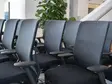Reihe schwarzer Bürostühle mit Netzrücken und Armlehnen in einem Büro oder Konferenzraum, im Hintergrund große grüne Pflanze und Treppe