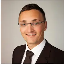 Heiser mit Brille, Hemd und Krawatte vor hellem, neutralem Hintergrund.