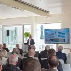 grusswort-bert-sutter-neubau-event.jpg Bert Sutter spricht vor Publikum bei einer Veranstaltung zum Neubau der Sutter Medizintechnik GmbH, im Hintergrund ist ein Bildschirm mit der Visualisierung des neuen Gebäudes zu sehen.