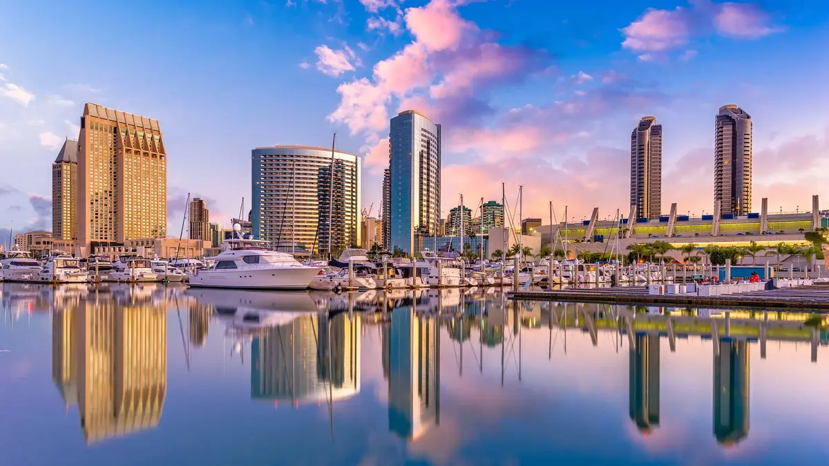 Panoramic view of San Diego skyline at sunset with high-rise buildings, yachts in the marina, and colorful sky.