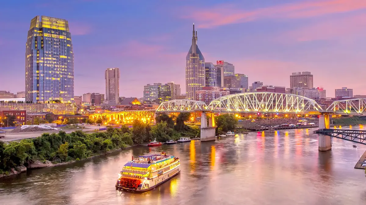 Nashville skyline at dusk with Cumberland River, illuminated bridge, riverboat, and downtown skyscrapers.
