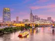 Nashville skyline at dusk with Cumberland River, illuminated bridge, riverboat, and downtown skyscrapers.
