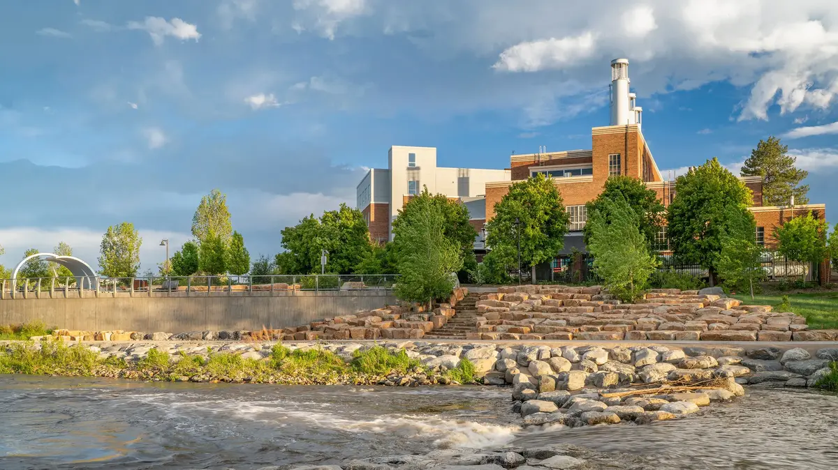 University building beside a river with stone embankment, trees, and a cloudy sky.