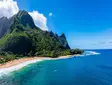 Tropical coastal landscape on Kauai, Hawaii, featuring steep lush green mountains, a sandy beach, and turquoise ocean under a blue sky