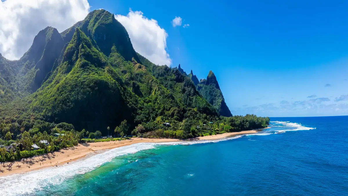 Tropical coastal landscape on Kauai, Hawaii, featuring steep lush green mountains, a sandy beach, and turquoise ocean under a blue sky