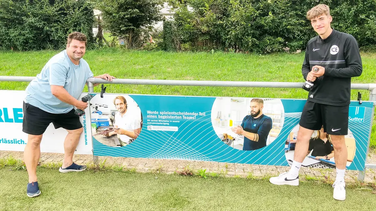 Zwei Männer montieren eine Sutter-Werbebande mit Karriereaufruf am Fußballplatz des FC Emmendingen, beide halten einen Akkuschrauber in der Hand und lächeln.