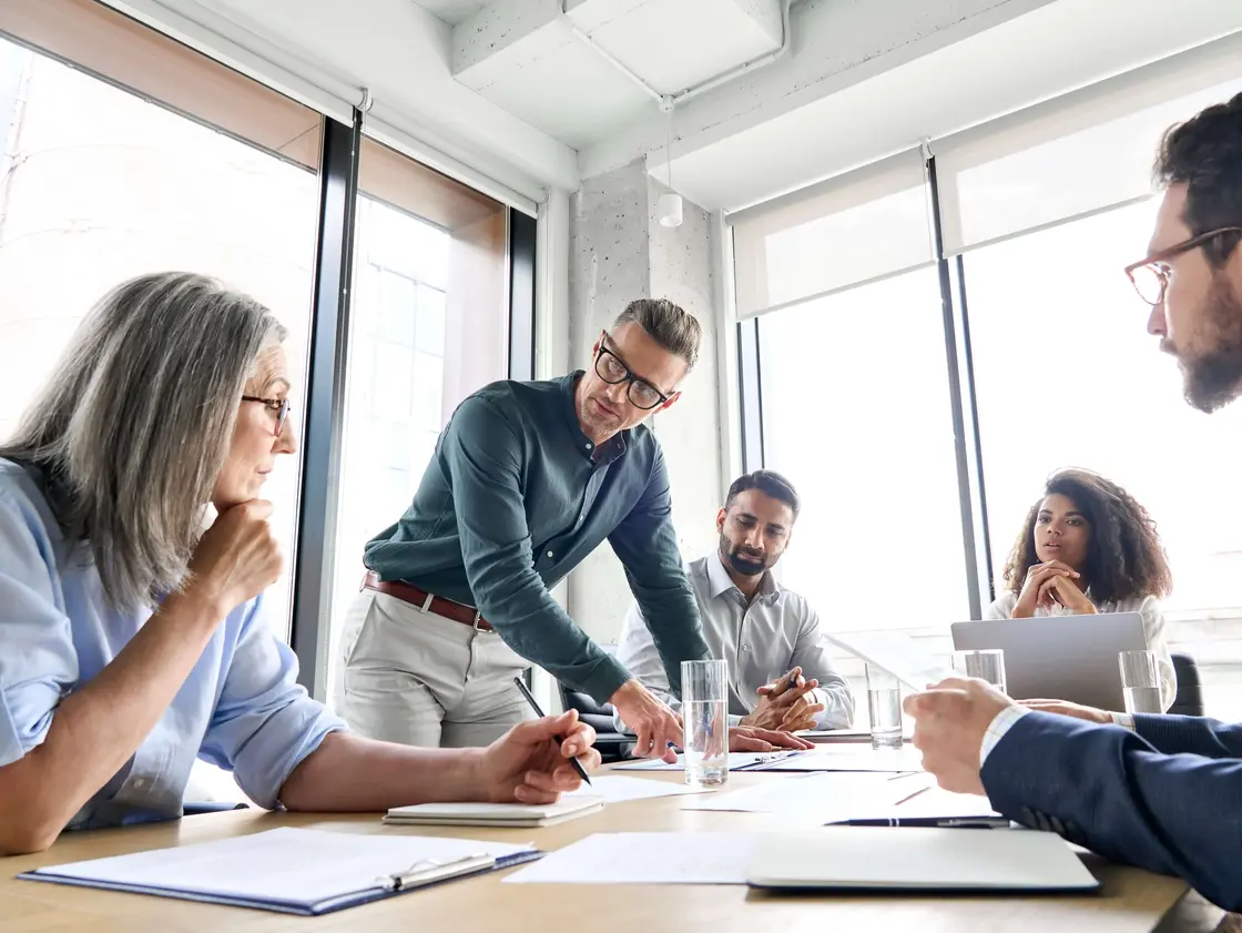 Besprechung mit fünf Personen in modernem Büro – Diskussion über Dokumente am Tisch mit Notizbüchern und Wassergläsern, geeignet für medizinisches Projektmanagement oder Teamarbeit.