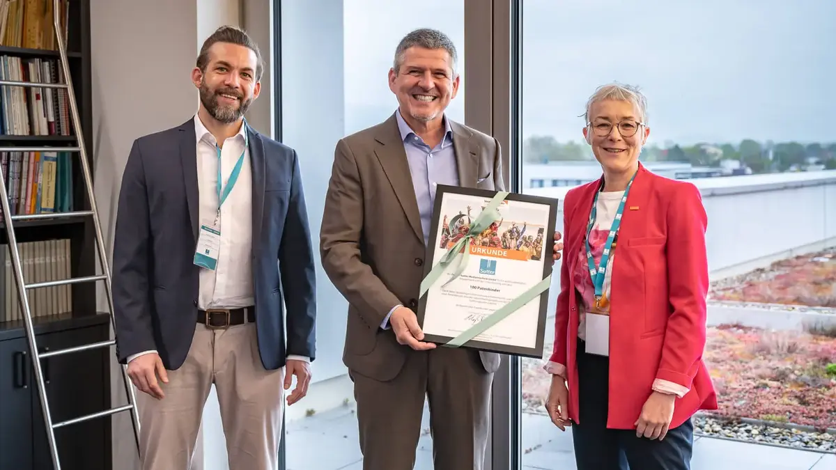 sutter-medizintechnik-world-vision-urkunde.jpg Drei Personen stehen lächelnd zusammen; in der Mitte wird eine gerahmte Urkunde von World Vision mit einer grünen Schleife überreicht, die 100 Patenschaften würdigt; im Hintergrund Büro mit Bücherregal und Fensterfront.