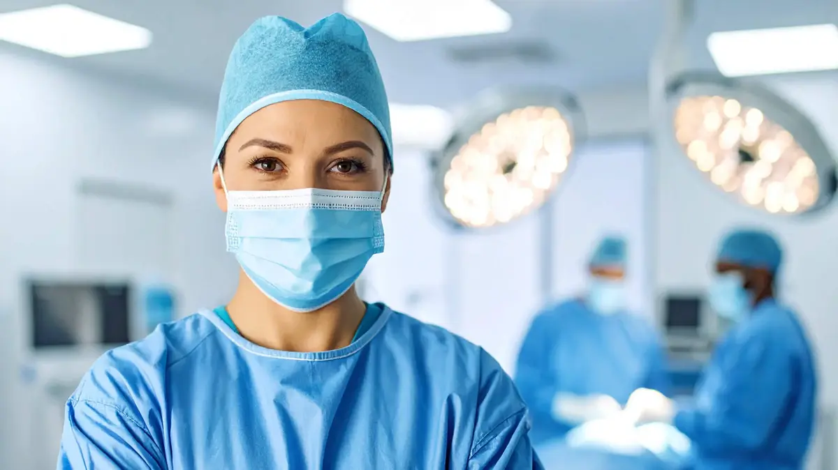 Junge Chirurgin mit OP-Maske und Kittel steht im Operationssaal, im Hintergrund arbeiten Kolleg:innen am OP-Tisch.