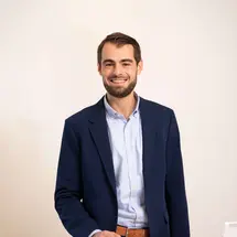 Person in blauem Blazer und hellblauem Hemd hinter weißem Tresen mit geöffnetem Laptop und Broschüren, Gesicht unscharf dargestellt