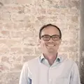 Person in hellblauem Hemd lächelnd vor einer Backsteinwand
