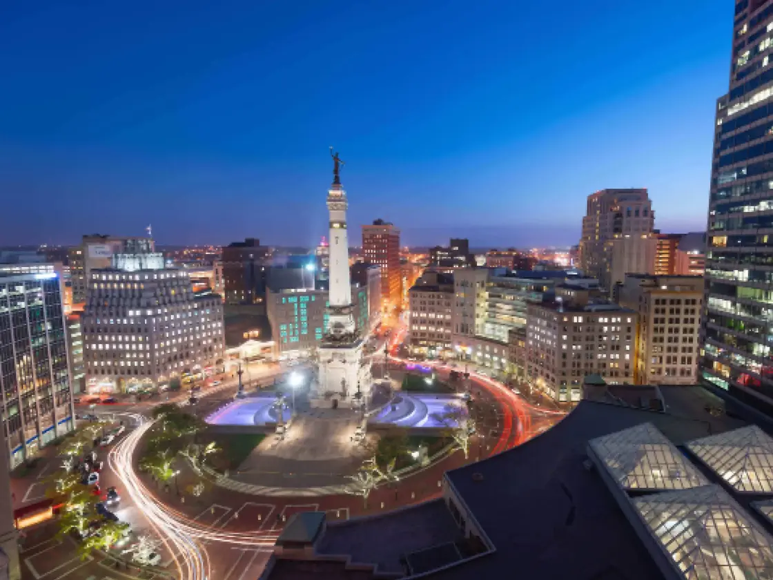 Aerial view of a city at dusk featuring an illuminated monument, surrounding buildings, and light trails from moving vehicles